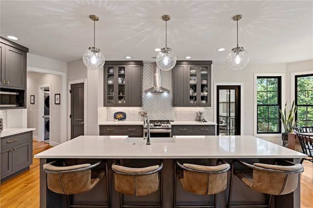 kitchen with hanging light fixtures, a kitchen island with sink, and wall chimney range hood