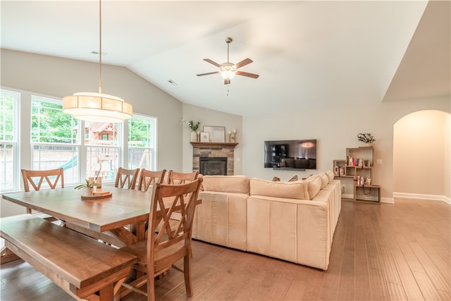 dining area featuring ceiling fan, a stone fireplace, light wood-type flooring, and vaulted ceiling