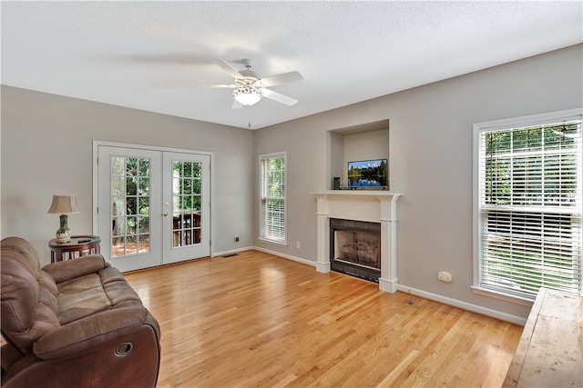 living room with a tiled fireplace, light wood-type flooring, a textured ceiling, ceiling fan, and french doors