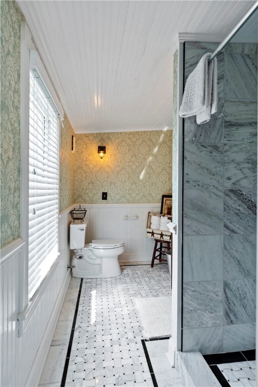 full bathroom with a wainscoted wall, a tile shower, toilet, and wallpapered walls