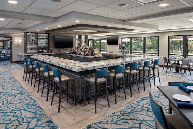 bar with light hardwood / wood-style floors, a healthy amount of sunlight, light stone countertops, and a paneled ceiling