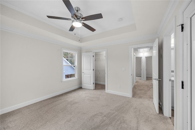 unfurnished bedroom with a spacious closet, a tray ceiling, ceiling fan, crown molding, and light colored carpet