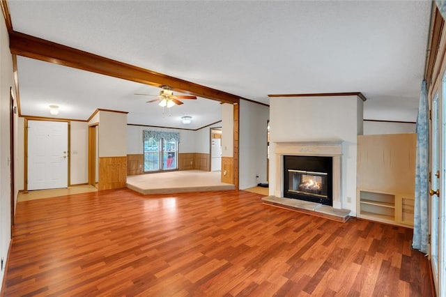 unfurnished living room featuring lofted ceiling with beams, a wainscoted wall, wood finished floors, a glass covered fireplace, and crown molding