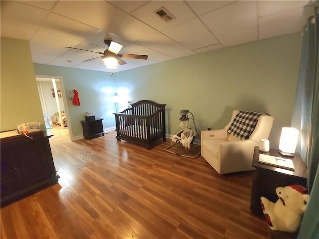 bedroom with hardwood / wood-style floors, a nursery area, a drop ceiling, and ceiling fan