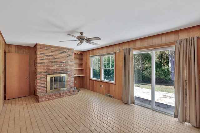 unfurnished living room with a fireplace, built in shelves, ceiling fan, and wooden walls