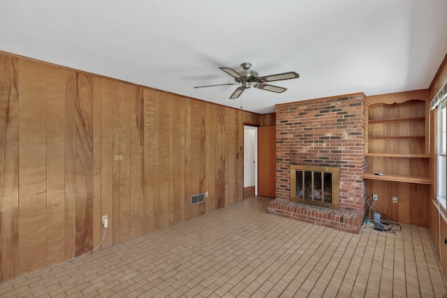 unfurnished living room with built in shelves, a brick fireplace, ceiling fan, and wooden walls