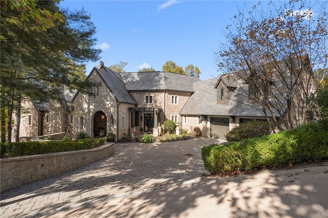 french country style house with a garage, stone siding, decorative driveway, a chimney, and a high end roof