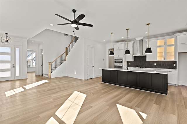 kitchen with white cabinets, a center island with sink, hanging light fixtures, and wall chimney range hood