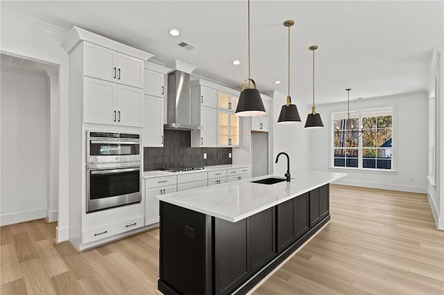 kitchen with sink, wall chimney exhaust hood, hanging light fixtures, an island with sink, and white cabinets
