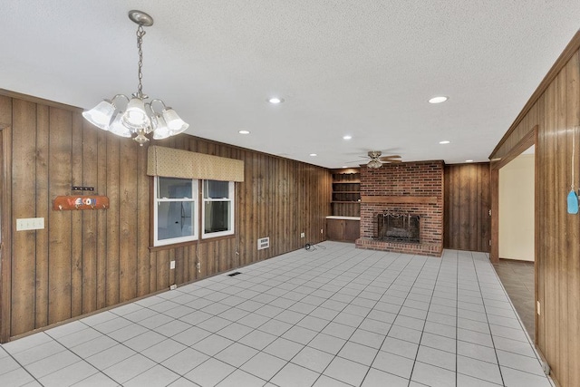 unfurnished living room featuring a fireplace, ceiling fan with notable chandelier, wooden walls, and light tile patterned flooring