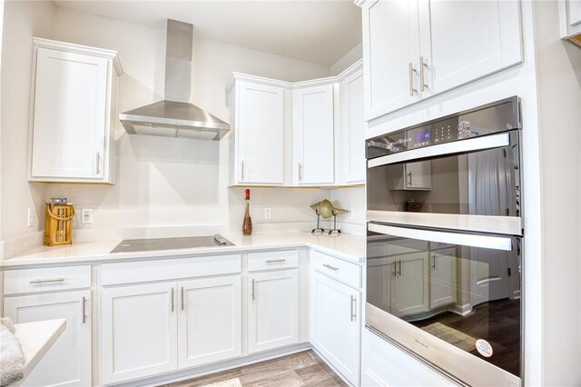 kitchen with light hardwood / wood-style flooring, white cabinetry, black electric stovetop, double wall oven, and wall chimney exhaust hood