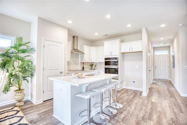 kitchen with wall chimney range hood, sink, white cabinetry, a kitchen island with sink, and black appliances