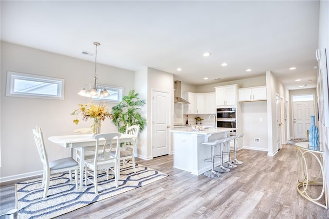 kitchen with decorative light fixtures, white cabinets, a kitchen island with sink, light wood-type flooring, and wall chimney exhaust hood