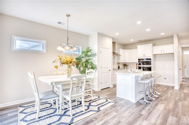 kitchen featuring wall chimney exhaust hood, white cabinetry, a center island with sink, light hardwood / wood-style flooring, and double oven