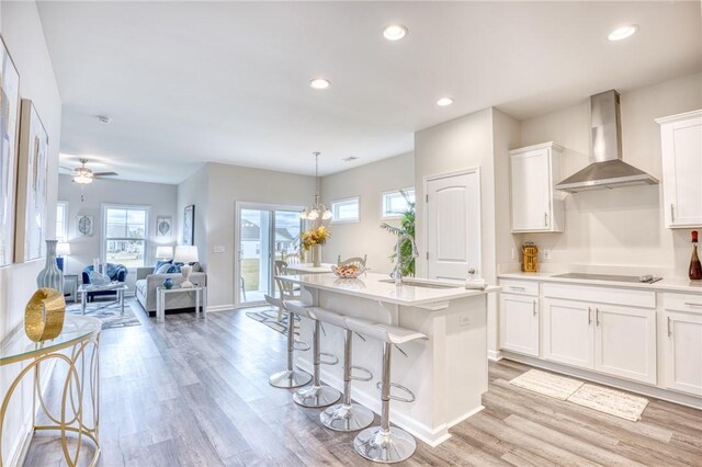 kitchen with light hardwood / wood-style floors, an island with sink, white cabinets, decorative light fixtures, and wall chimney exhaust hood