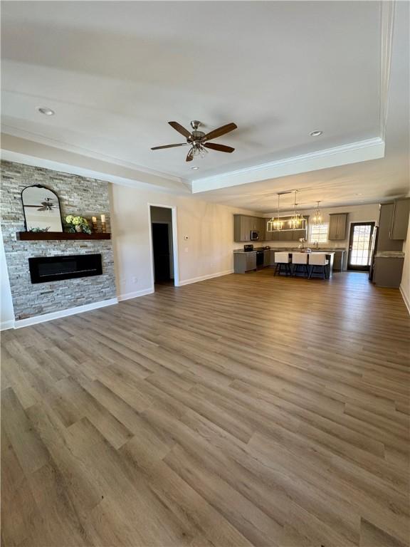 unfurnished living room featuring ceiling fan with notable chandelier, a tray ceiling, dark wood-type flooring, crown molding, and a stone fireplace