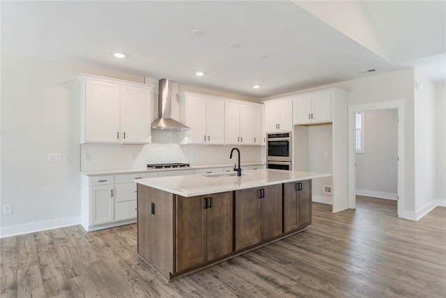 kitchen with wall chimney exhaust hood, white cabinetry, sink, and an island with sink