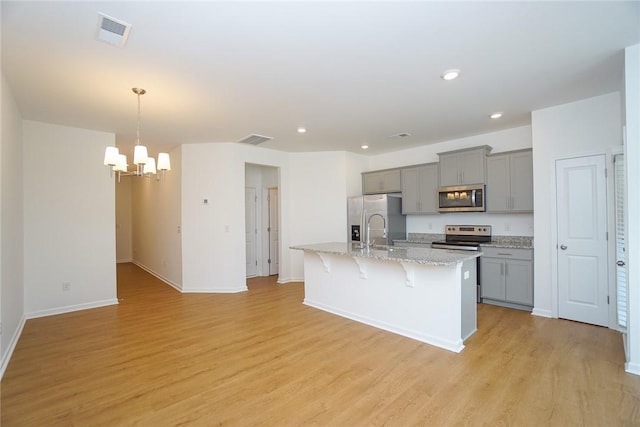 kitchen with light stone counters, stainless steel appliances, a center island with sink, a chandelier, and gray cabinets