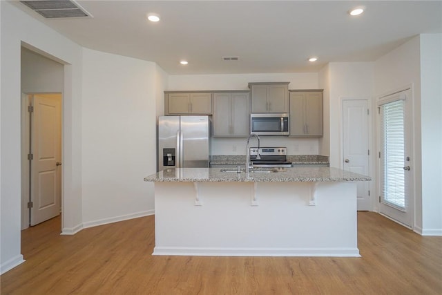 kitchen featuring gray cabinetry, a center island with sink, a kitchen breakfast bar, light stone counters, and stainless steel appliances