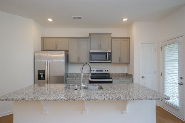 kitchen with a center island with sink, sink, light stone countertops, appliances with stainless steel finishes, and a kitchen bar