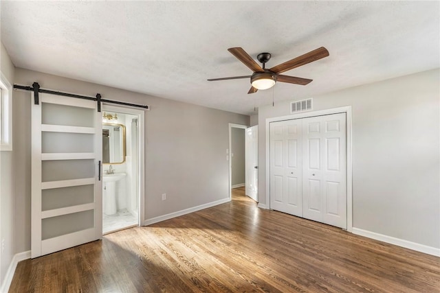 unfurnished bedroom featuring ceiling fan, a barn door, hardwood / wood-style flooring, connected bathroom, and a closet