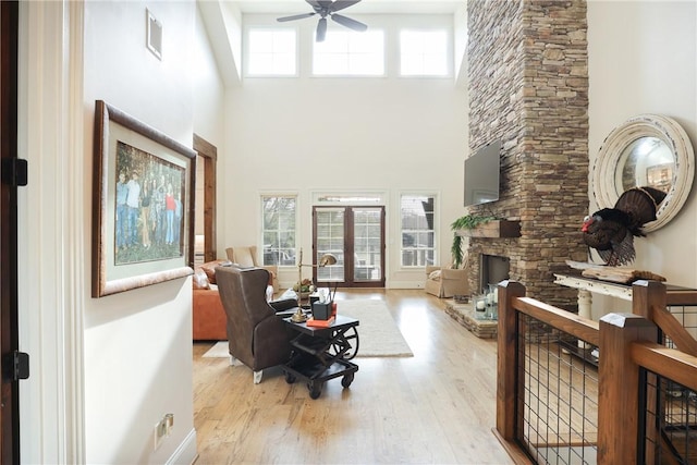 living room with french doors, ceiling fan, light hardwood / wood-style flooring, a high ceiling, and a stone fireplace