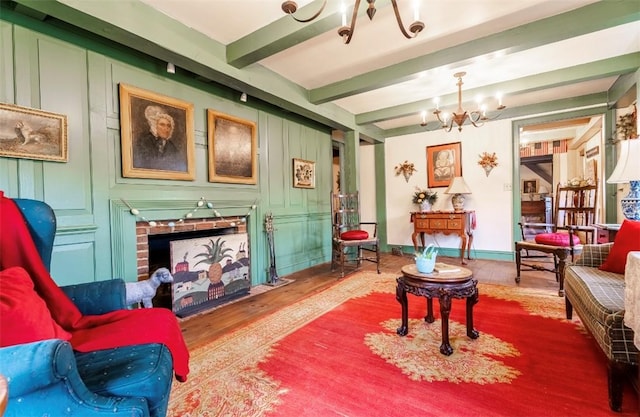 living area with wood finished floors, beamed ceiling, a brick fireplace, baseboards, and a notable chandelier