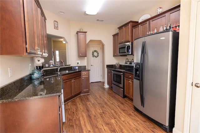 kitchen with dark wood-type flooring, a sink, dark stone countertops, arched walkways, and appliances with stainless steel finishes