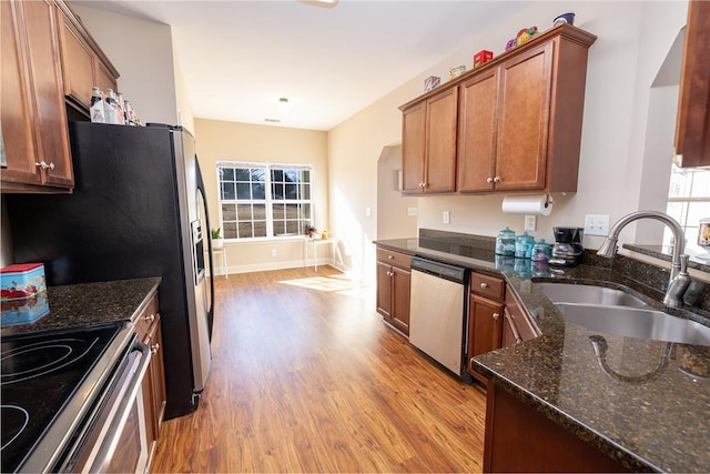 kitchen with brown cabinets, a sink, wood finished floors, stainless steel appliances, and dark stone counters