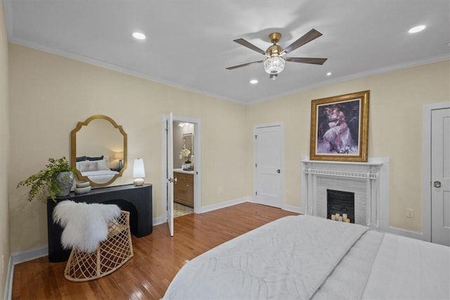 bedroom featuring hardwood / wood-style floors, a fireplace, ceiling fan, ensuite bathroom, and crown molding