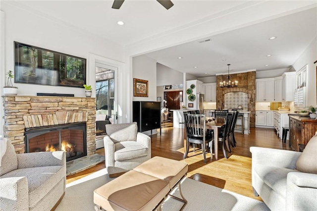 living room featuring ceiling fan with notable chandelier, hardwood / wood-style floors, beam ceiling, and a stone fireplace