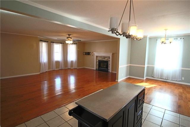 unfurnished living room with light tile patterned flooring, ornamental molding, ceiling fan with notable chandelier, and a stone fireplace