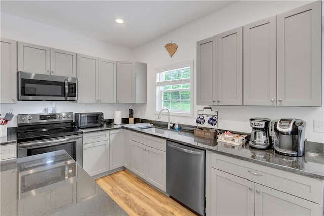 kitchen with appliances with stainless steel finishes, gray cabinetry, sink, light wood-type flooring, and dark stone counters