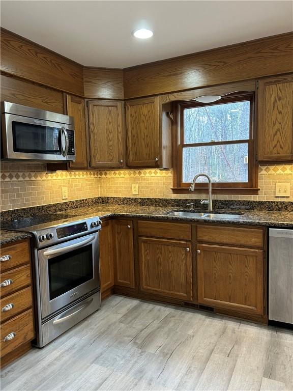 kitchen with light wood-style flooring, a sink, appliances with stainless steel finishes, backsplash, and dark stone countertops