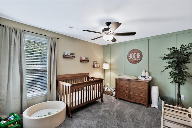 bedroom with a nursery area, ceiling fan, and dark carpet