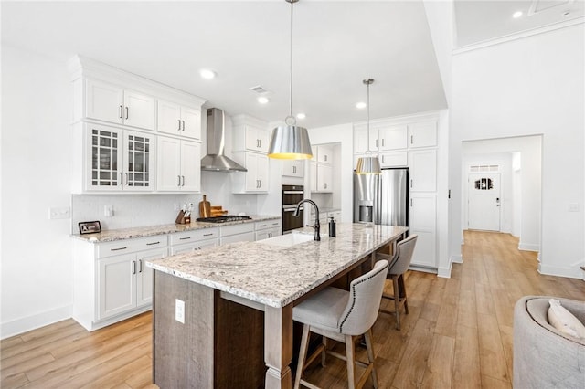 kitchen with pendant lighting, appliances with stainless steel finishes, white cabinetry, a center island with sink, and wall chimney exhaust hood