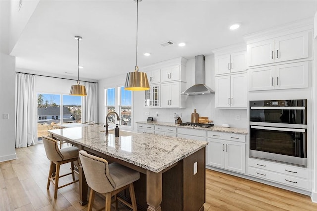 kitchen featuring white cabinetry, decorative light fixtures, double oven, an island with sink, and wall chimney range hood