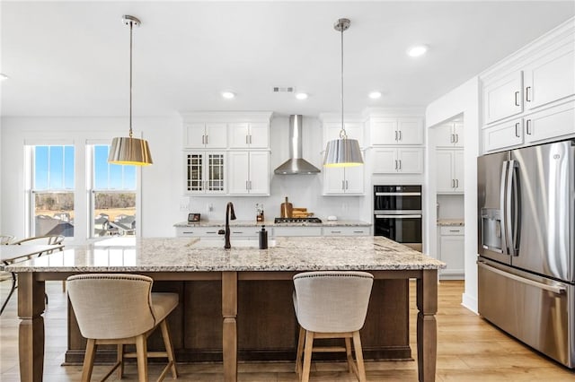 kitchen featuring wall chimney exhaust hood, stainless steel appliances, an island with sink, and hanging light fixtures