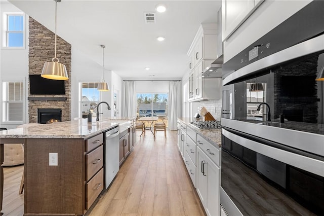 kitchen featuring appliances with stainless steel finishes, light stone countertops, white cabinets, a center island with sink, and a stone fireplace