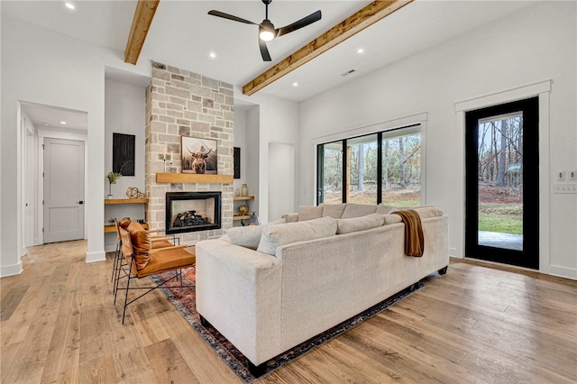 living room featuring a stone fireplace, a towering ceiling, beamed ceiling, ceiling fan, and light wood-type flooring