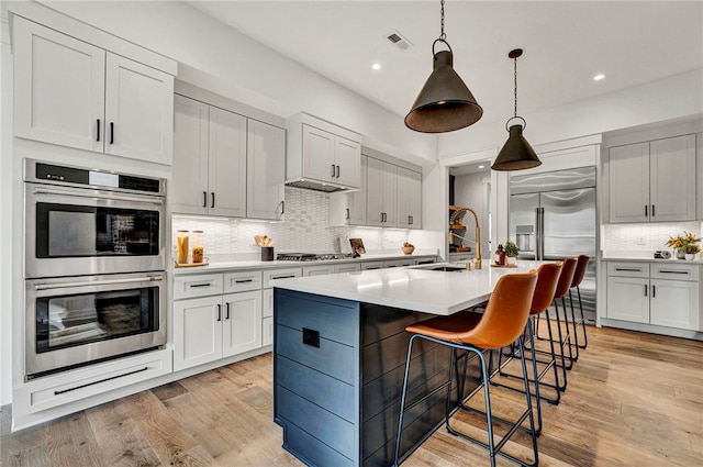 kitchen featuring stainless steel appliances, light hardwood / wood-style flooring, a center island with sink, and decorative light fixtures