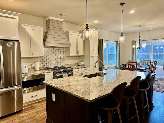 kitchen with sink, appliances with stainless steel finishes, an island with sink, custom range hood, and white cabinets