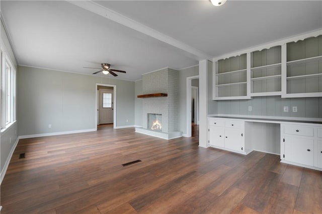 unfurnished office featuring dark wood-type flooring, built in desk, a brick fireplace, ornamental molding, and ceiling fan