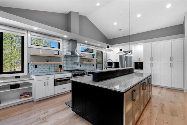 kitchen featuring decorative light fixtures, white cabinetry, an island with sink, light stone counters, and wall chimney exhaust hood