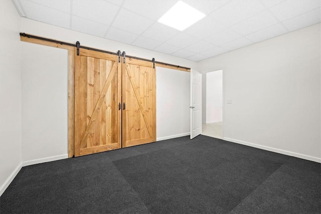 empty room with a paneled ceiling, a barn door, dark carpet, and baseboards