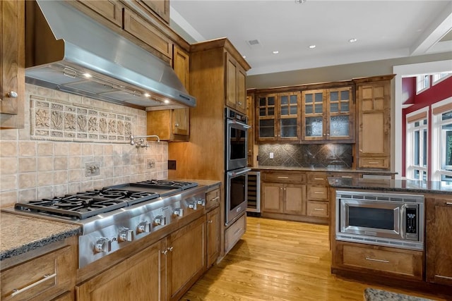 kitchen featuring appliances with stainless steel finishes, stone countertops, ventilation hood, backsplash, and light hardwood / wood-style floors