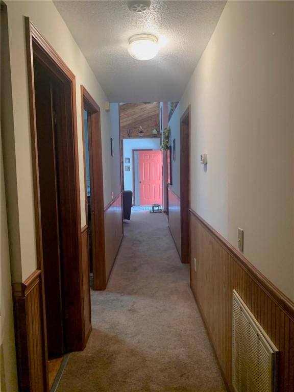 corridor with wooden walls, wainscoting, a textured ceiling, and light colored carpet