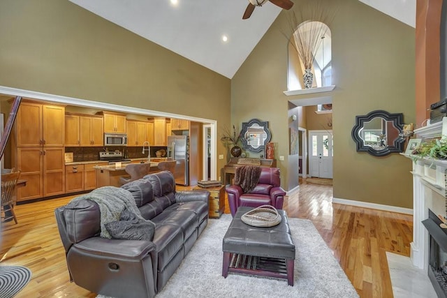 living area featuring baseboards, a ceiling fan, a fireplace with flush hearth, light wood-style floors, and high vaulted ceiling