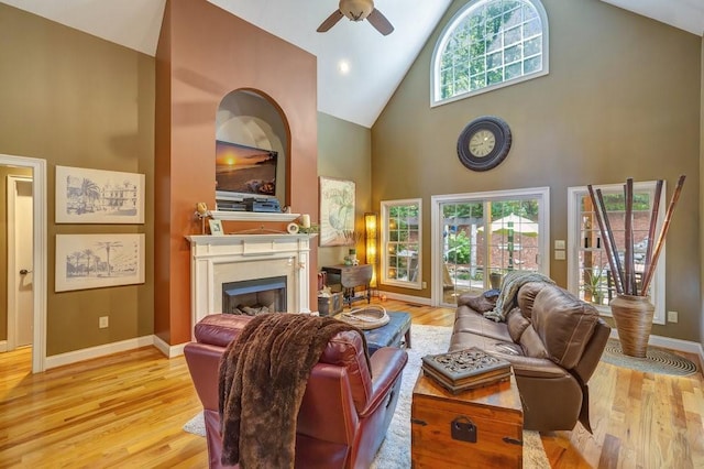 living room with baseboards, ceiling fan, light wood-style floors, a fireplace, and high vaulted ceiling