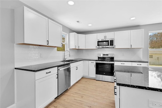 kitchen featuring light wood finished floors, white cabinets, dark stone countertops, stainless steel appliances, and a sink